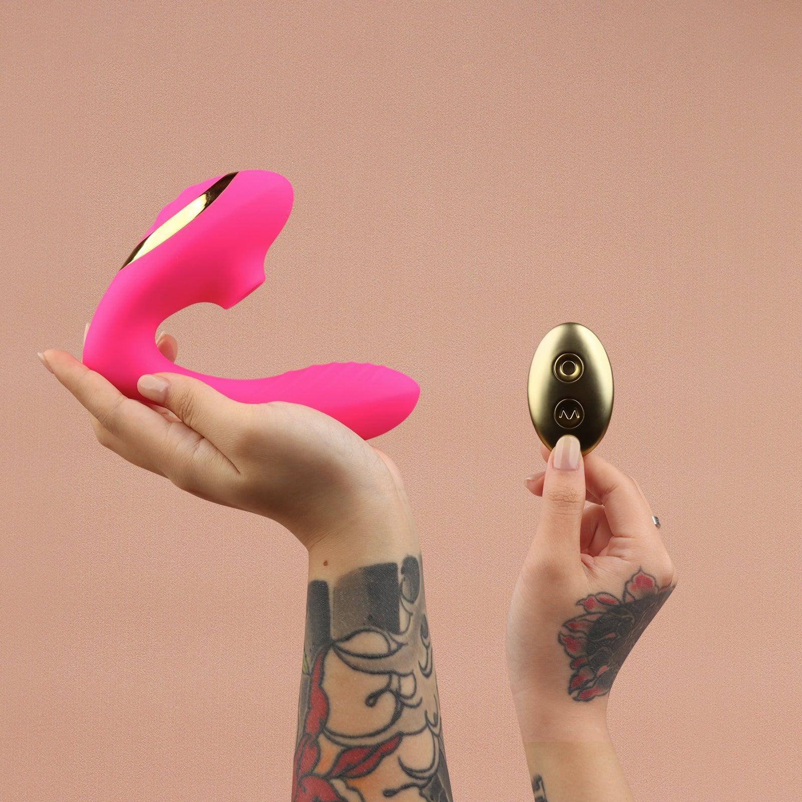 Pink OG 2 clitoral sucking vibrator with gold accents held in hand next to gold remote, peach background.